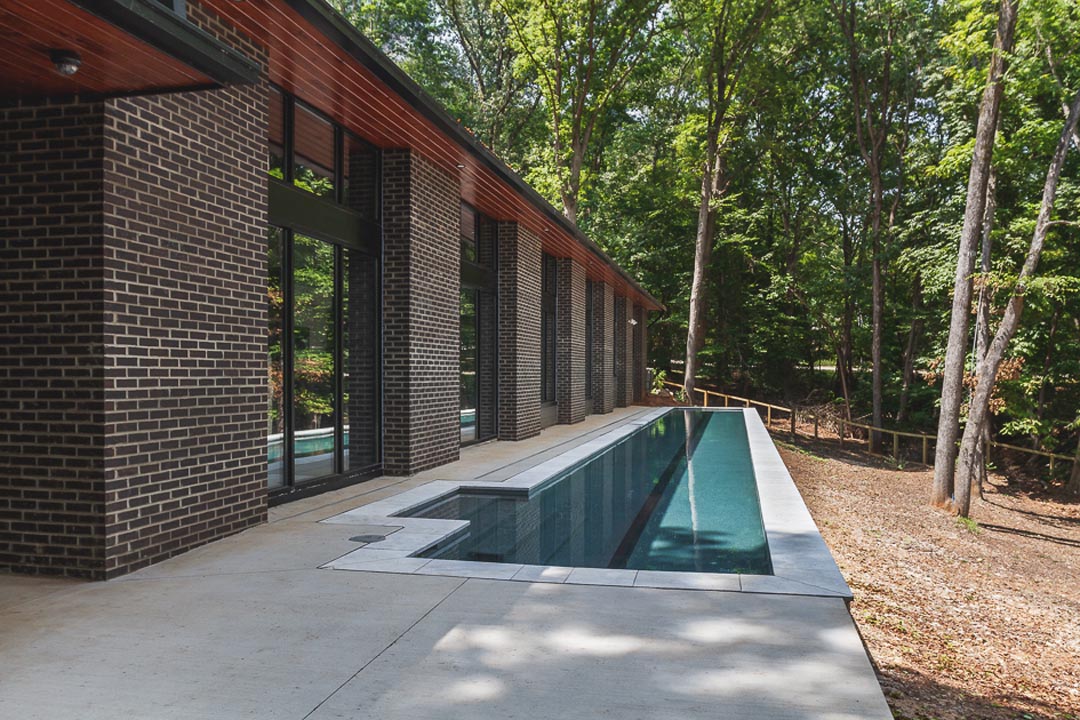 Narrow, elongated outdoor swimming pool next to a modern brick building with large windows, surrounded by trees and natural landscape.