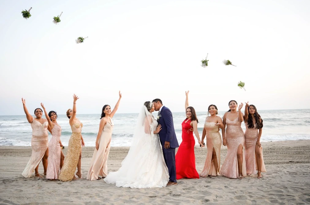 Bride and groom kissing on beach while bridesmaids throw bouquets in the air; American wedding traditions.