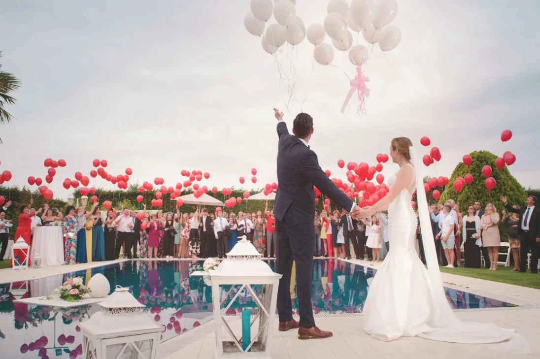 Man in tuxedo and women in white wedding gown celebrating by releasing balloons over a pool; American wedding traditions.