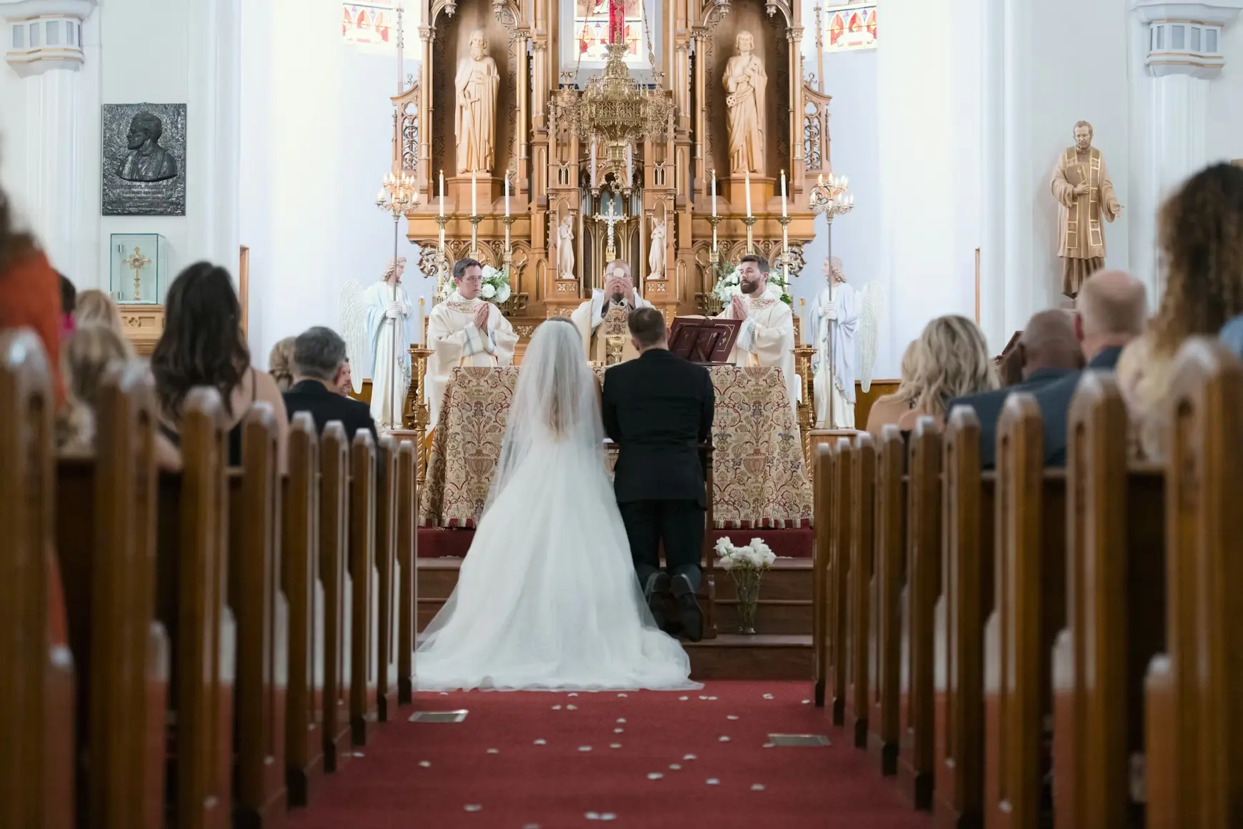 Bride and groom kneeling in front of alter while the priest marries the couple in front of guests.