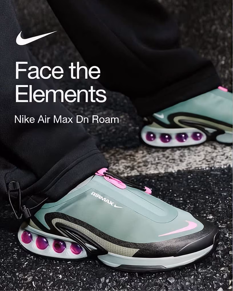 Close-up of Nike Air Max Dn Roam shoes in teal, black, and pink with visible air cushioning on the sole, worn with black pants on a textured pavement background, with text 'Face the Elements' and Nike logo.