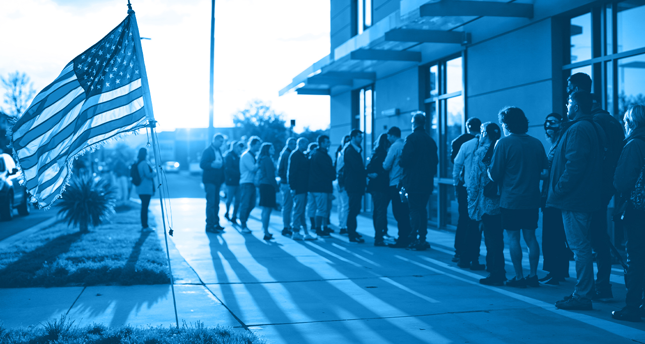 People standing in a line outside a building with a U.S. flag in the foreground, symbolizing immigration or government services in the United States.