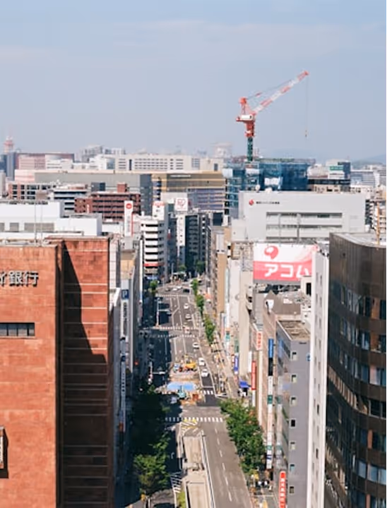 Urban cityscape featuring a street lined with buildings, a construction crane in the background, and clear sky overhead.
