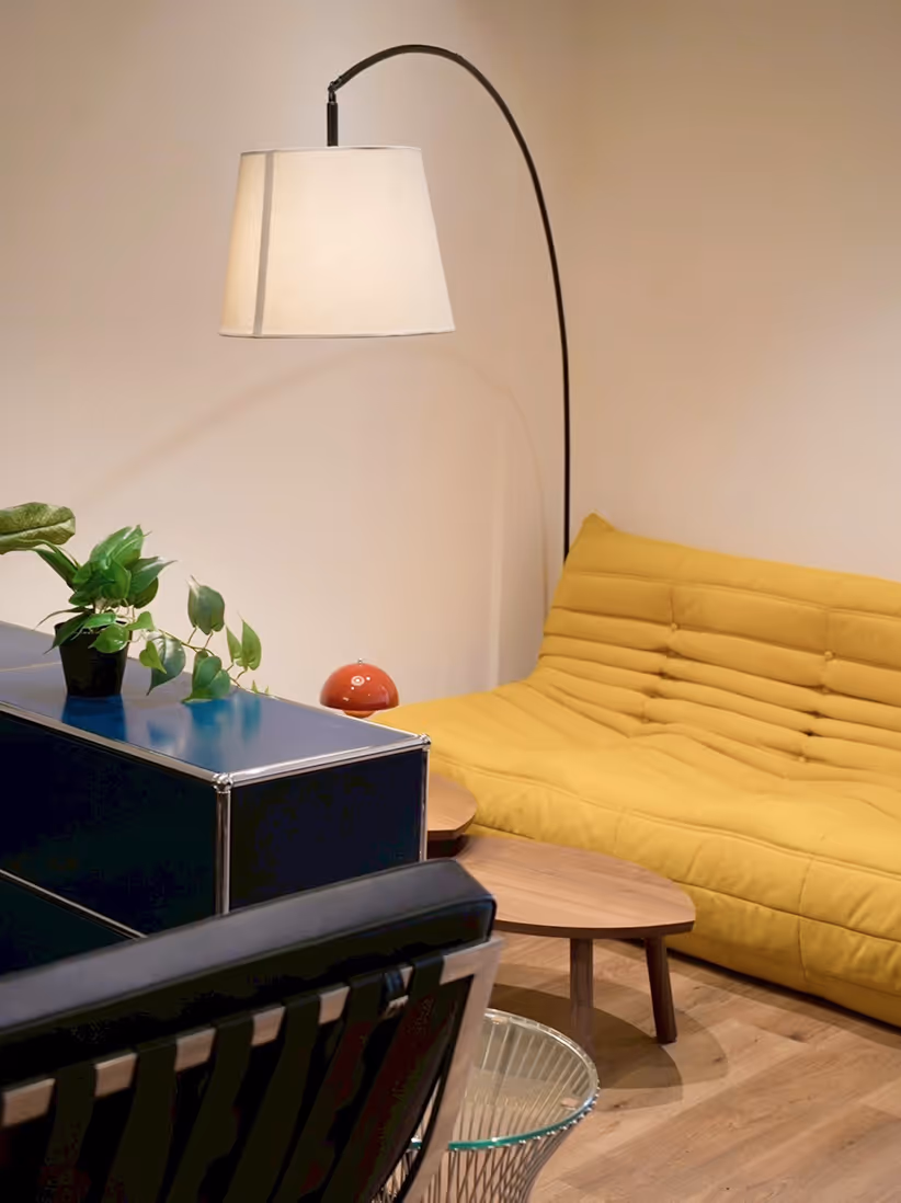Modern living room corner with a yellow cushioned sofa, an arched floor lamp with a white shade, a wooden coffee table, and a blue cabinet topped with a green potted plant.