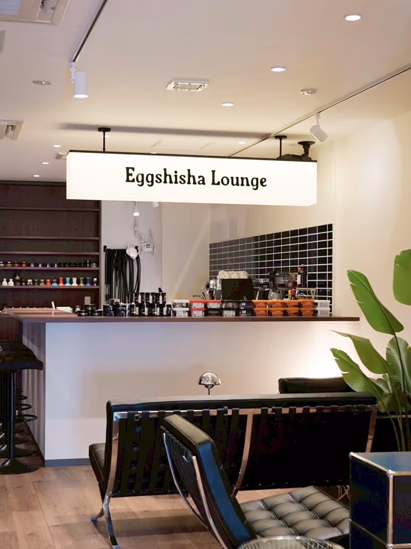 Modern lounge interior with a backlit sign reading 'Eggshisha Lounge' above a counter with various items, black seating, and a large green plant.