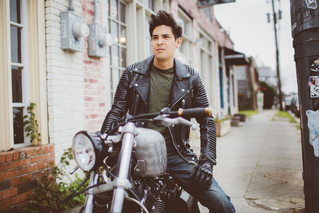 A man with black hair sits on his motorcycle looking ahead. He's parked on the sidewalk wearing a leather jacket with a green undershirt.