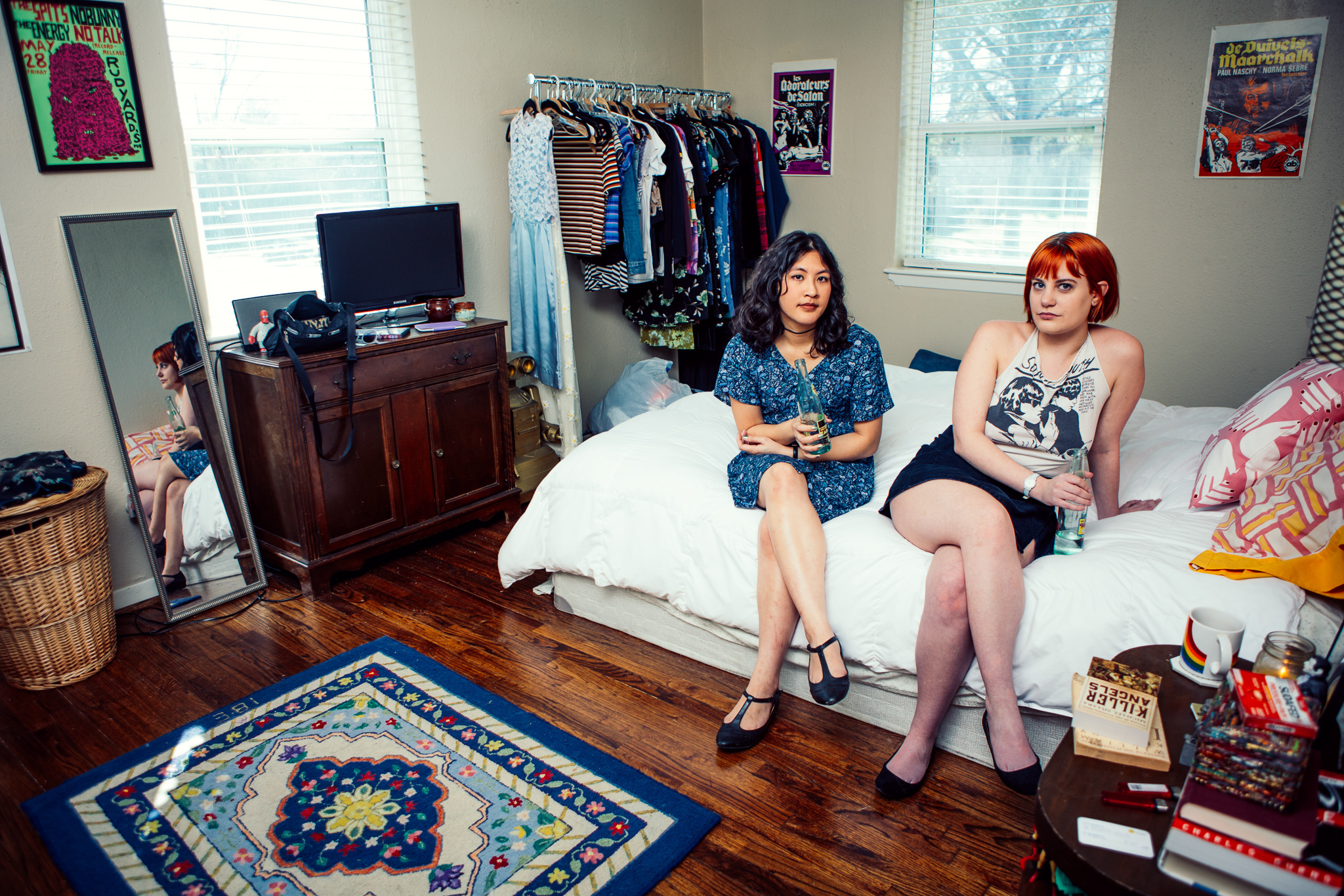 Two women sit on a bed with a white cover, and pink and orange pillows. They're sitting in some room with a clothing rack, and night stand, tv, mirror and rug.