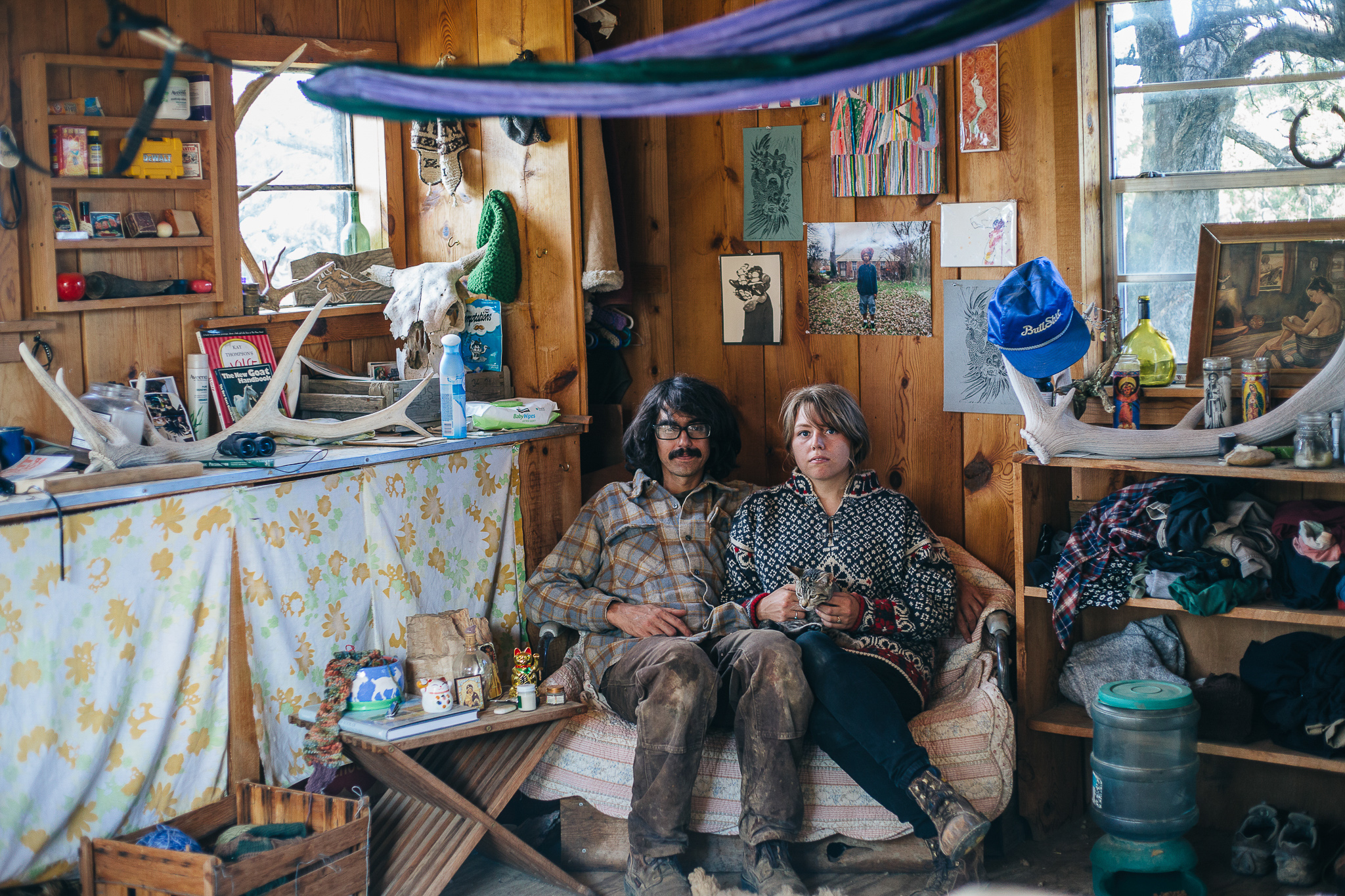 A couple sits on a small couch in a rustic cable with antlers adorning a window sill, a table, and a shelf. Small pieces of art are on the walls and a hammock hangs from the ceiling.
