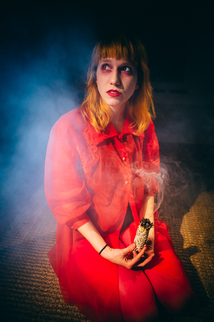 A red haired woman wearing all red with burgundy eye shadow is shown kneeling and holding burning sage while she looks off to the left.