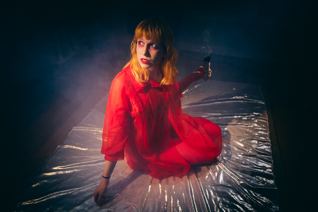 A woman in red holds sage as is burns. She is kneeling, legs together leaning and looking towards her right.