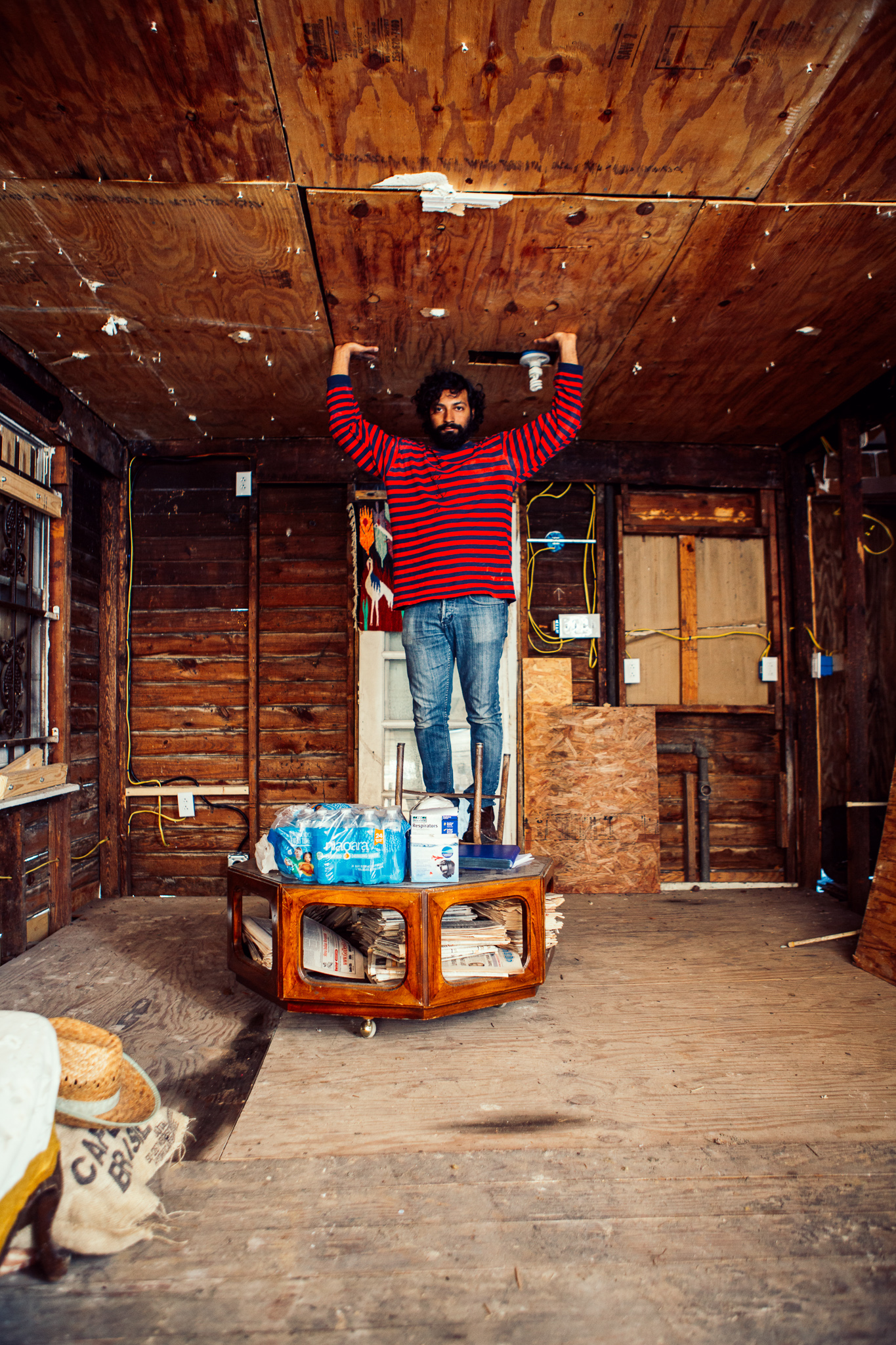 Inside a log cabin, a man in a red sweater stands on a table with his arms in the air 