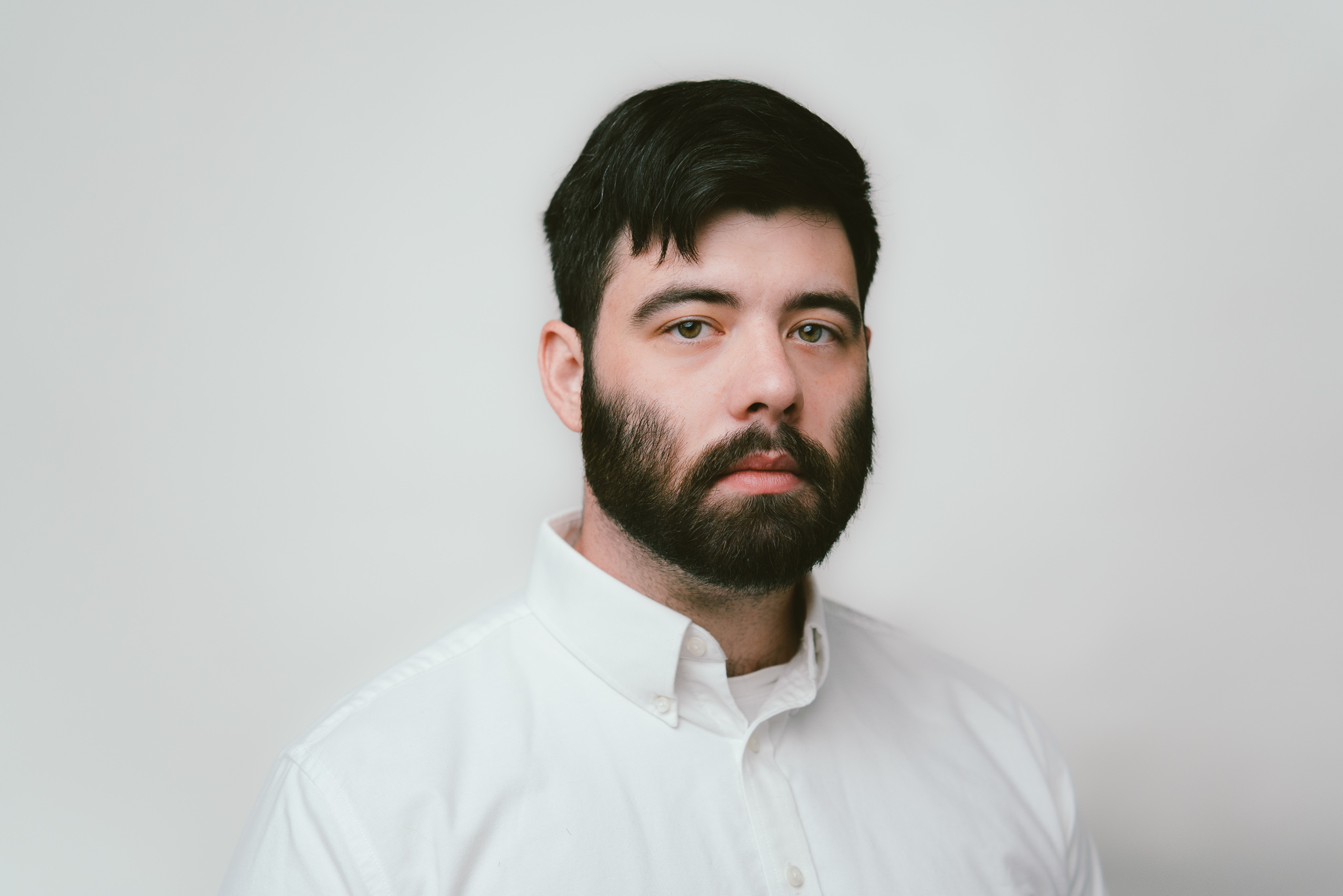 Portrait of a brunette haired man with a full beard on a white background wearing a white button down shirt