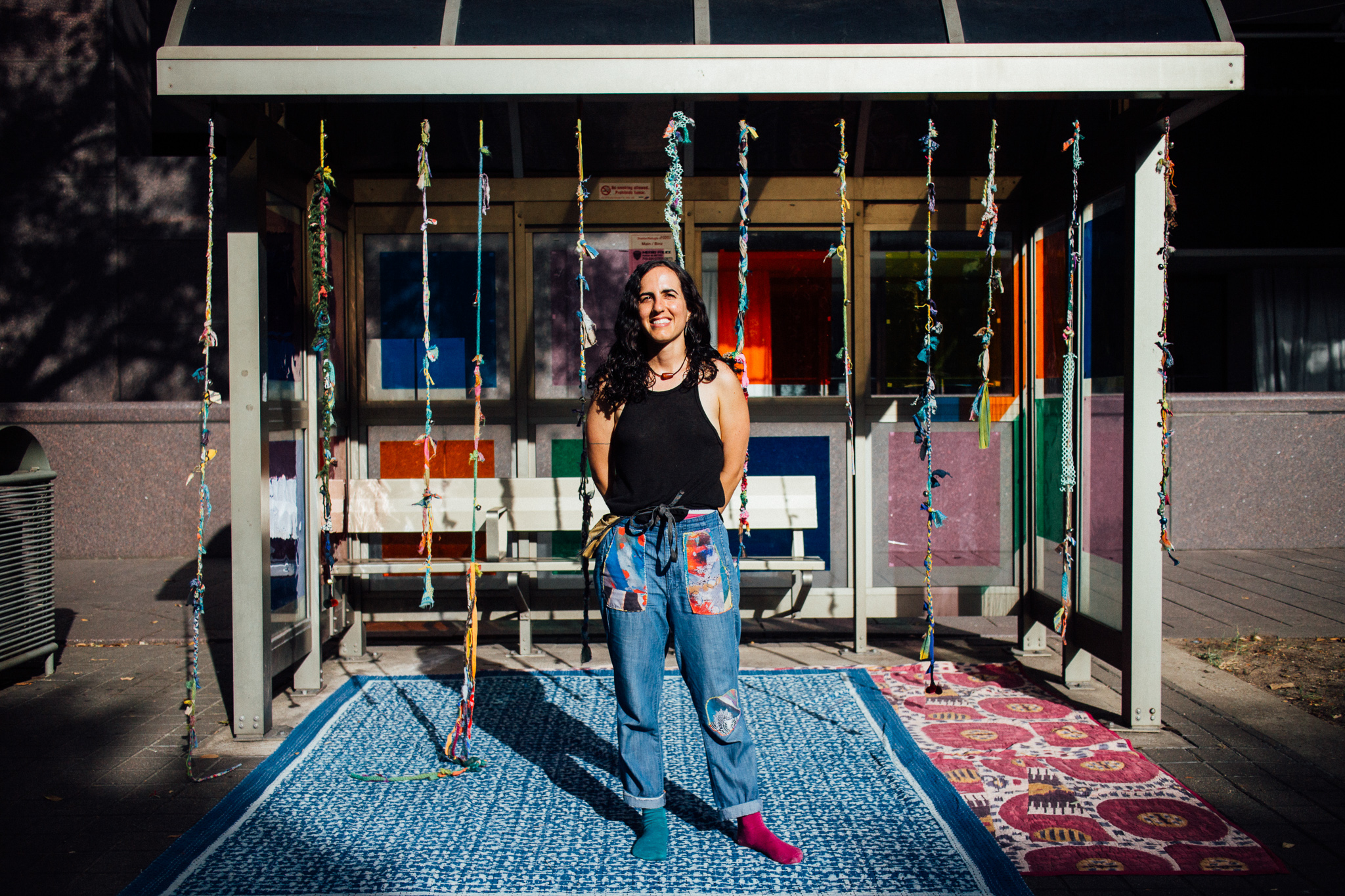 A woman wearing a black shirt and patch worked jeans stands in front of a bus stop decorated with hanging clothes patch and strung together