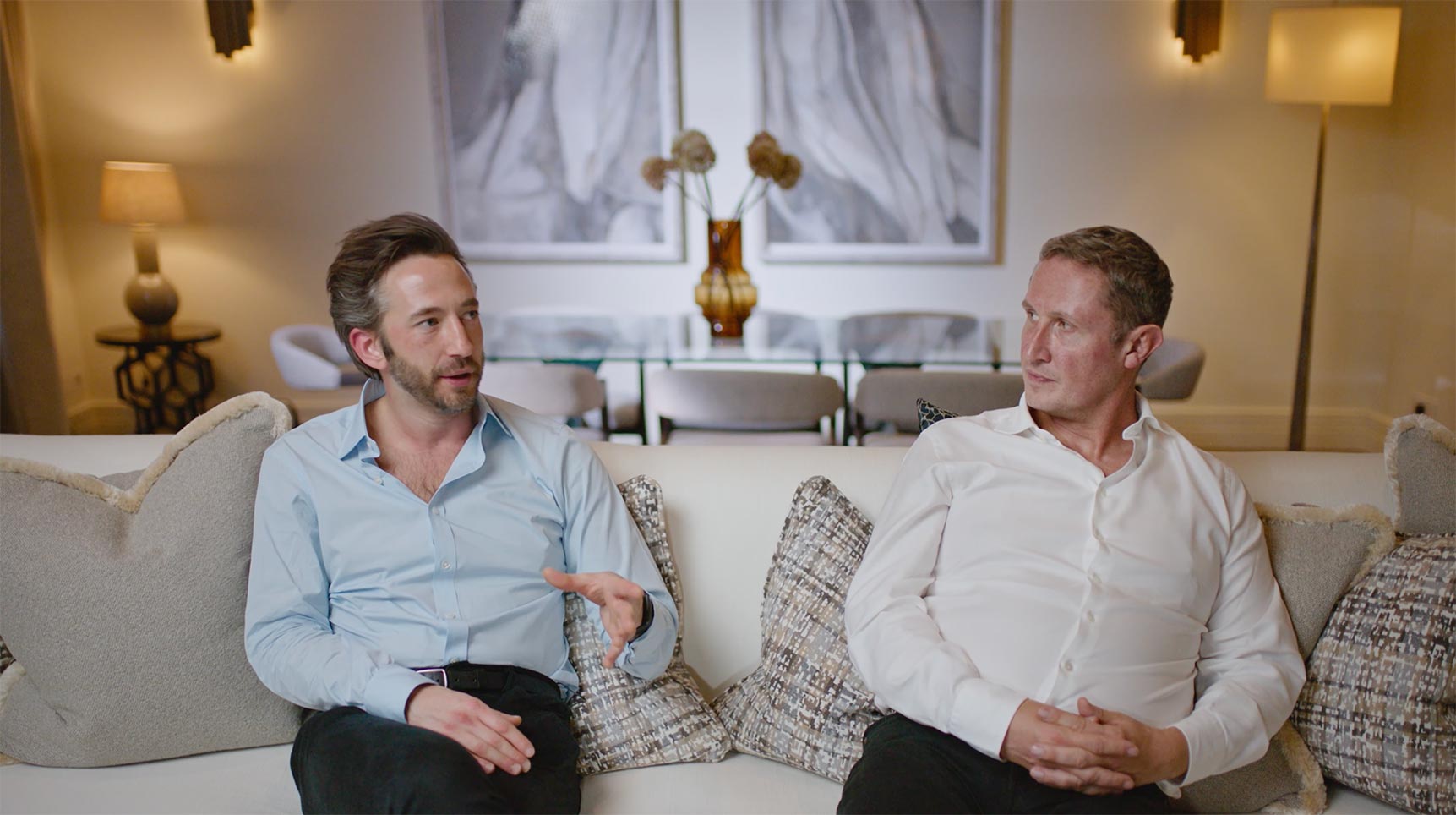 Two men sitting and talking on a couch in a modern living room with abstract art and warm lighting.
