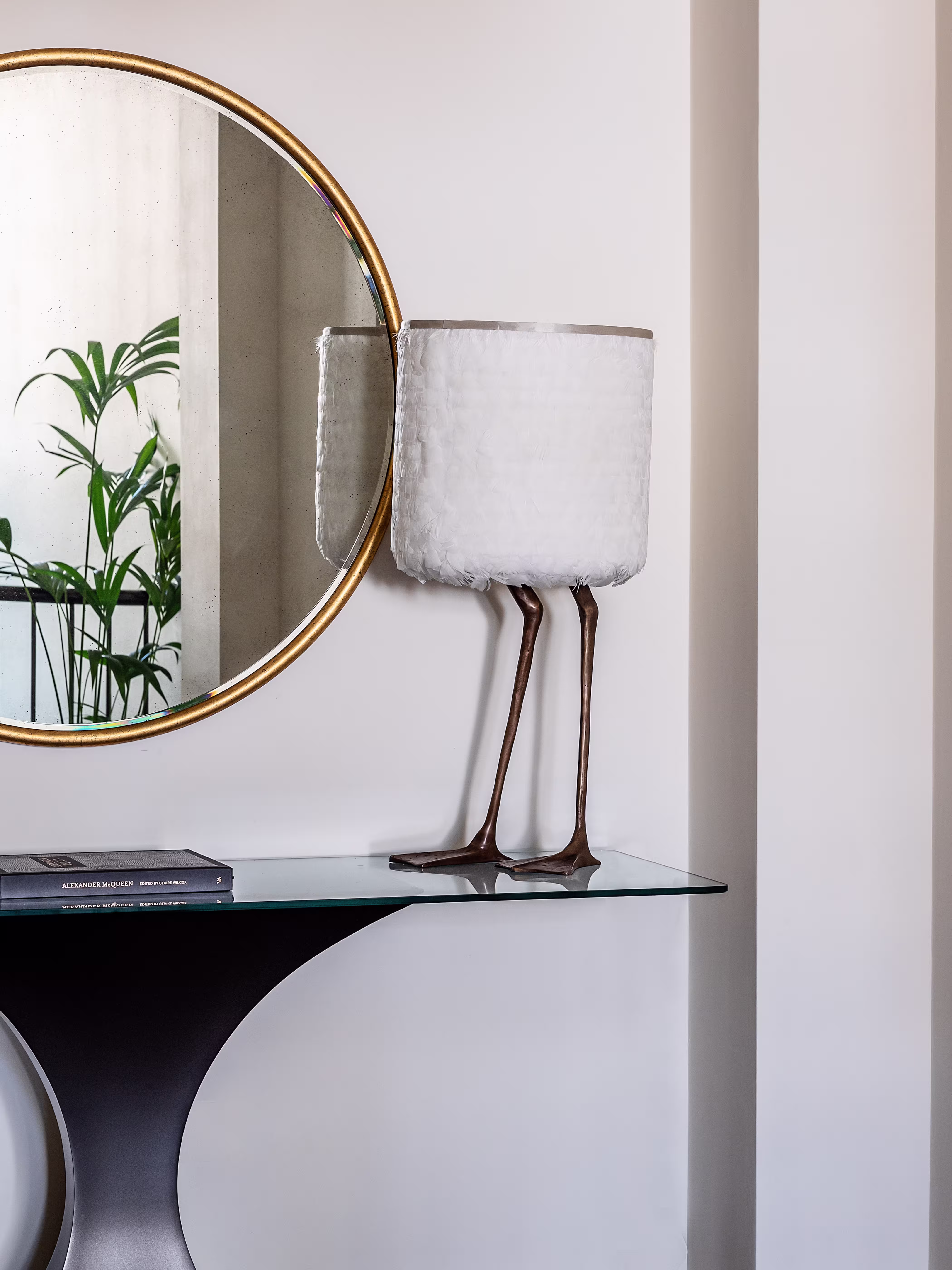 Decorative table lamp with white feathered shade and bronze bird-like legs on glass console table below round gold-framed mirror.