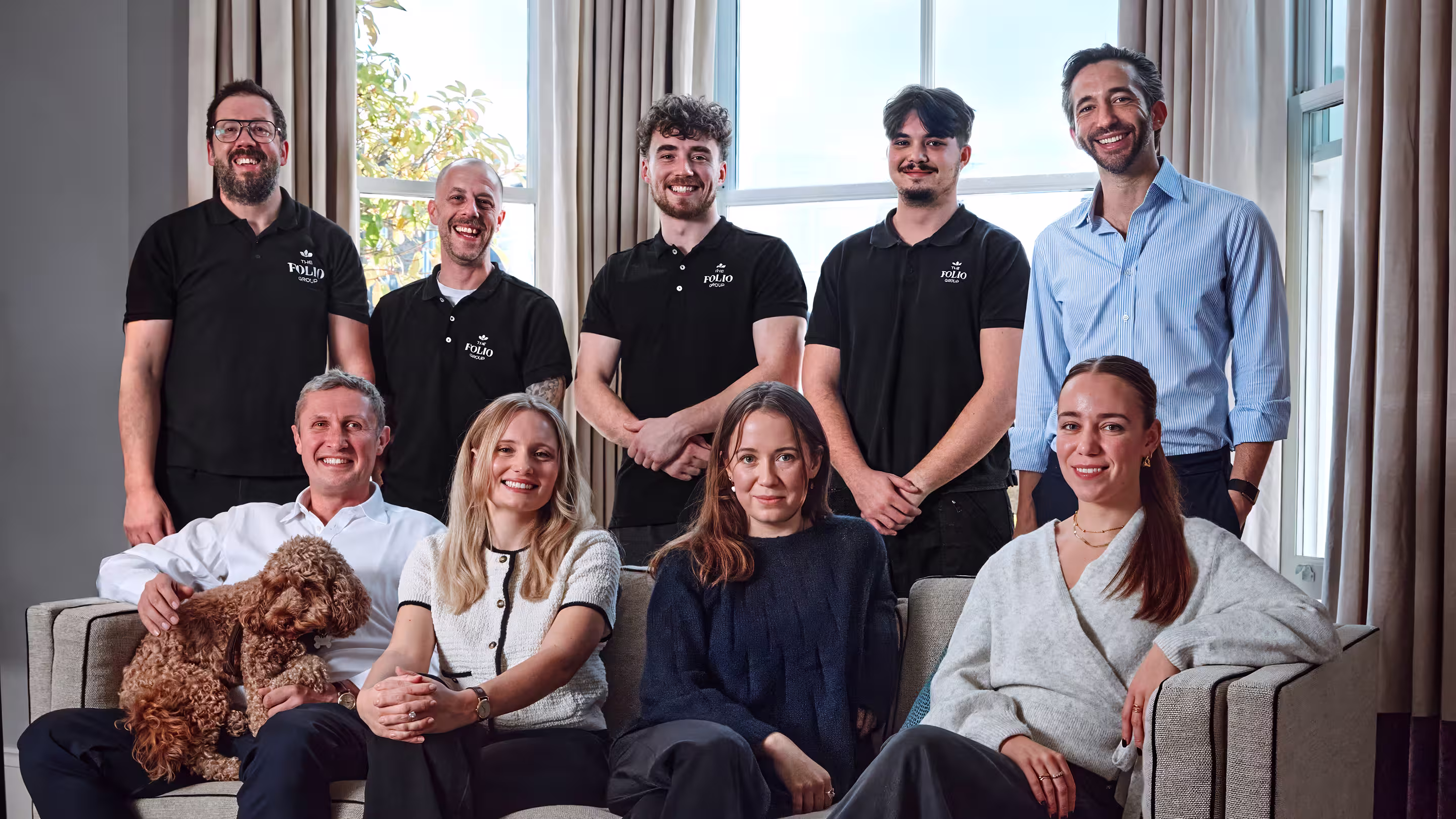 Group of nine smiling adults, four women sitting on a couch with a brown dog and five men standing behind, some wearing black Polo Group shirts.