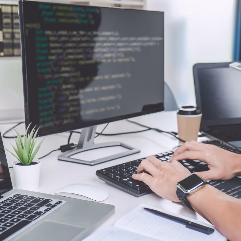 Personne tapant sur un clavier avec du code affiché sur un écran d'ordinateur, un ordinateur portable, une souris, un ordinateur portable avec un stylet, une petite plante et une tasse de café sur un bureau.