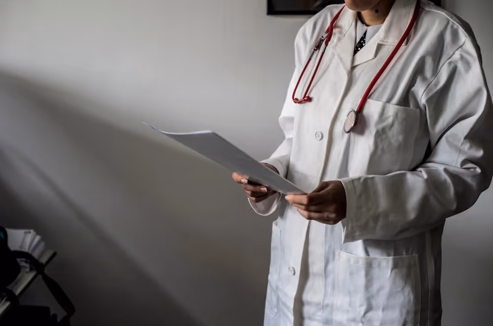 Professionnel de santé vêtu d'une blouse blanche et d'un stéthoscope rouge lisant un document à l'intérieur.