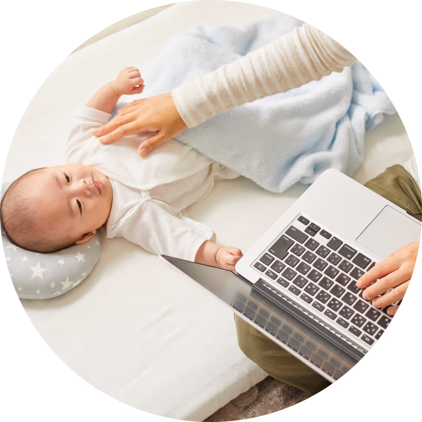 Baby lying on a cushion while an adult types on a laptop beside them with a hand gently placed on the baby.