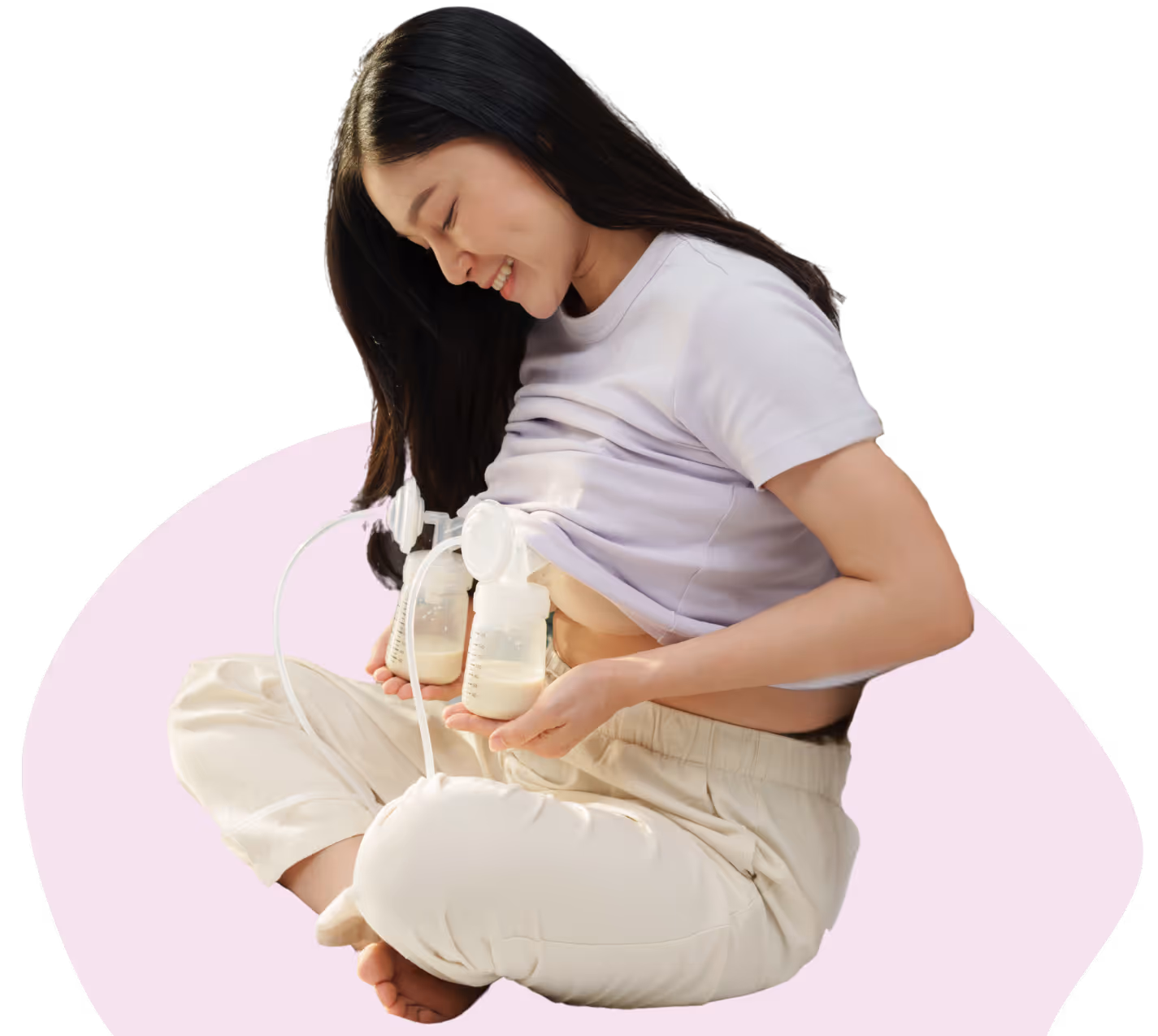 Smiling woman sitting cross-legged using a breast pump with milk bottles attached.