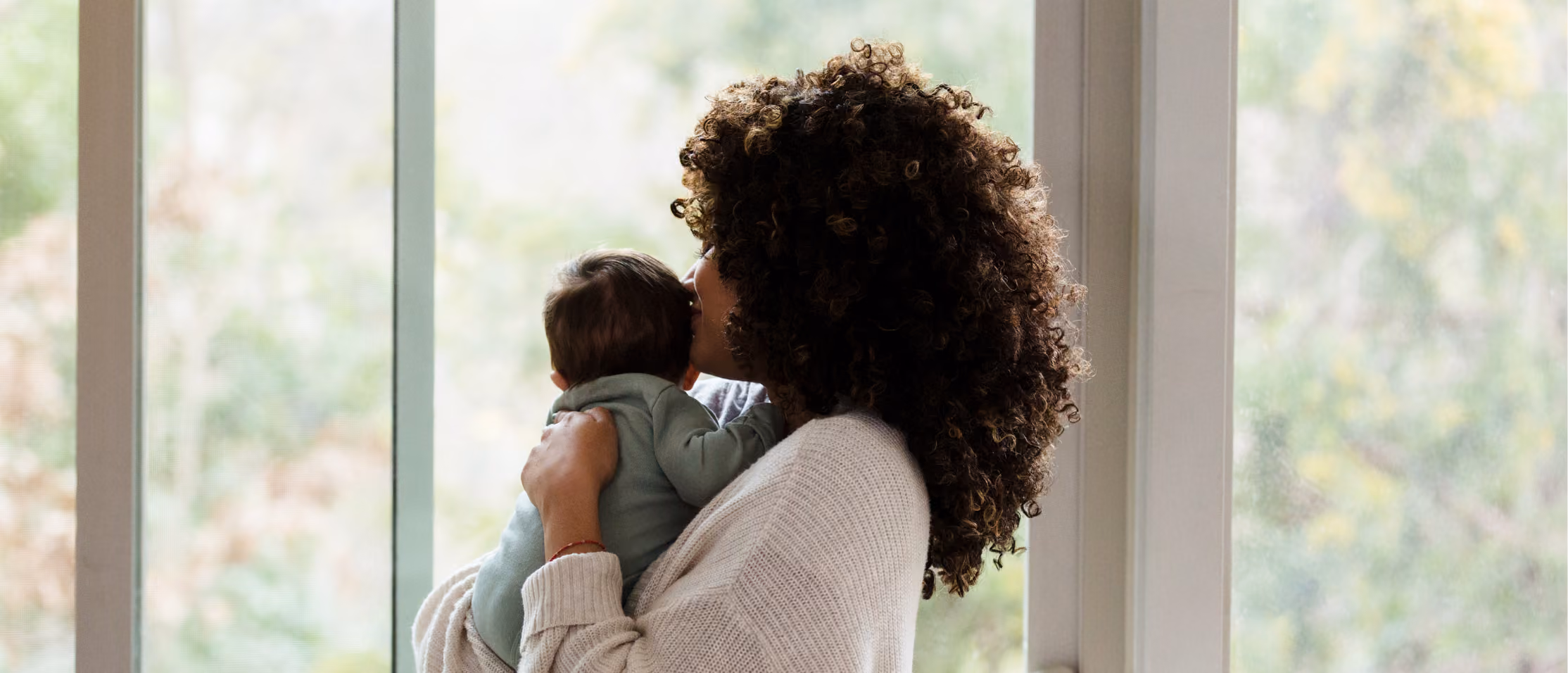 Woman with curly hair holding and kissing a baby near a large window with blurred outdoors view.