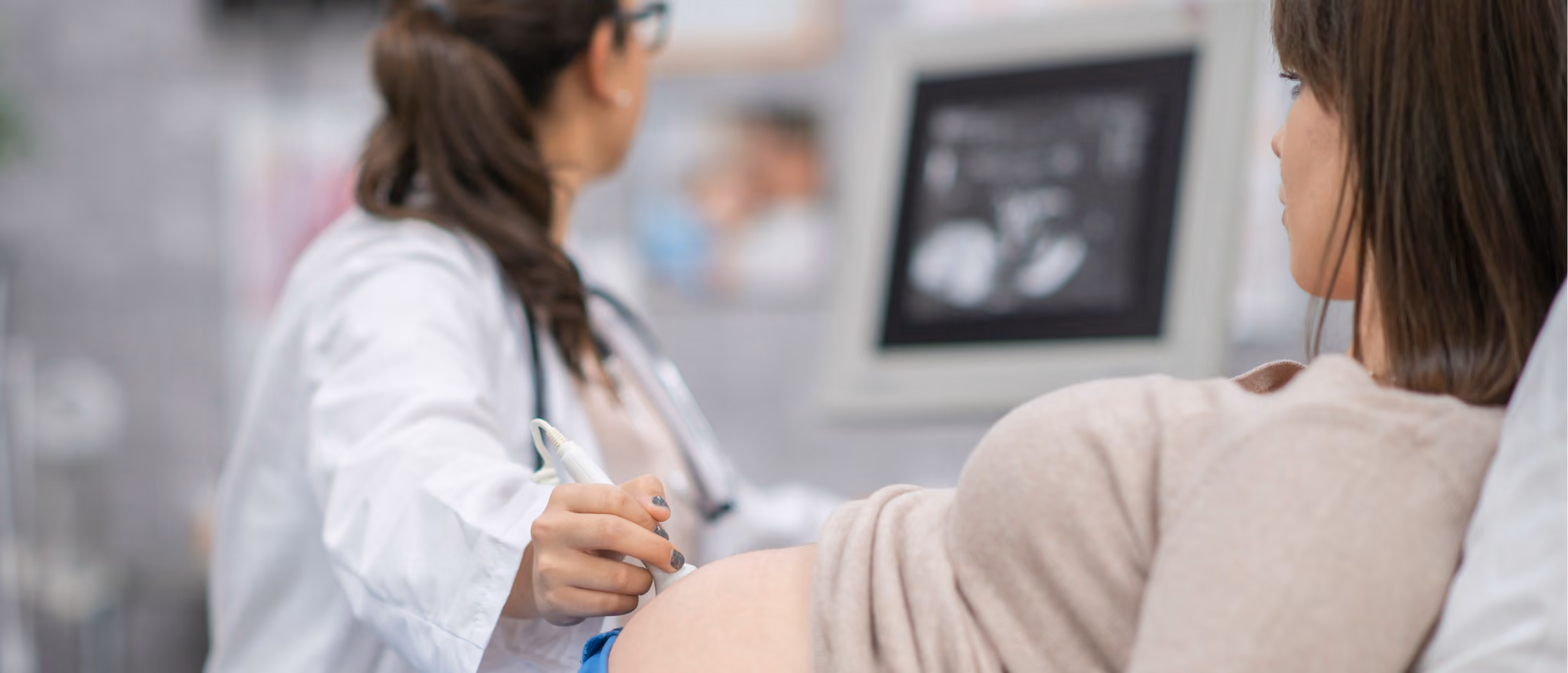 Doctor performing ultrasound on pregnant woman's belly with sonogram image in background.
