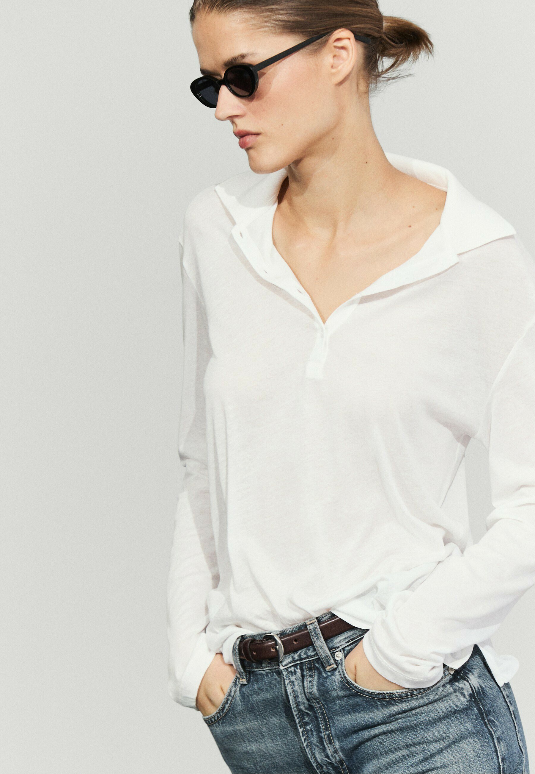 Donna con i capelli raccolti in uno chignon che indossa occhiali da sole neri, una camicia bianca a maniche lunghe con colletto aperto e jeans blu con le mani nelle tasche.