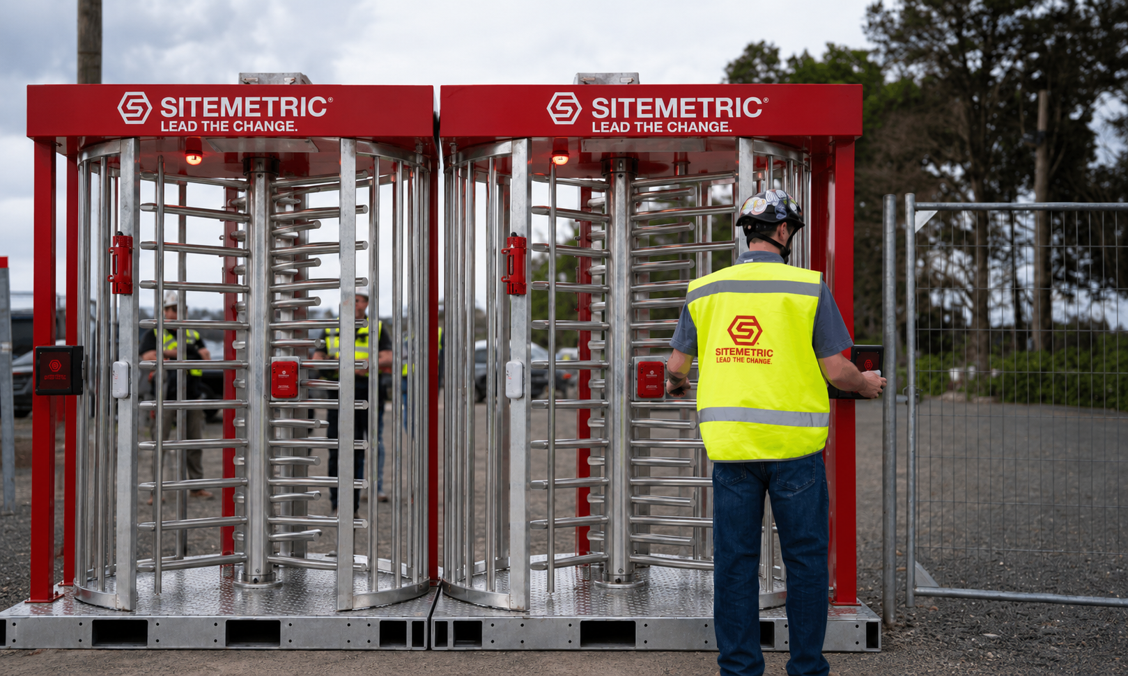A worker in a Sitemetric high-visibility vest badging in at a pair of Sitemetric Deluxe Turnstiles at a jobsite entrance.