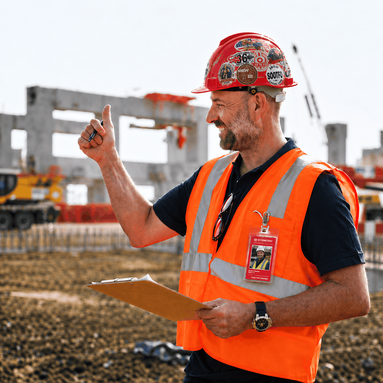 A smiling site manager in a red hard hat and orange high-visibility vest, with a Sitemetric photo badge clipped to the vest, giving a thumbs up on an active construction site.