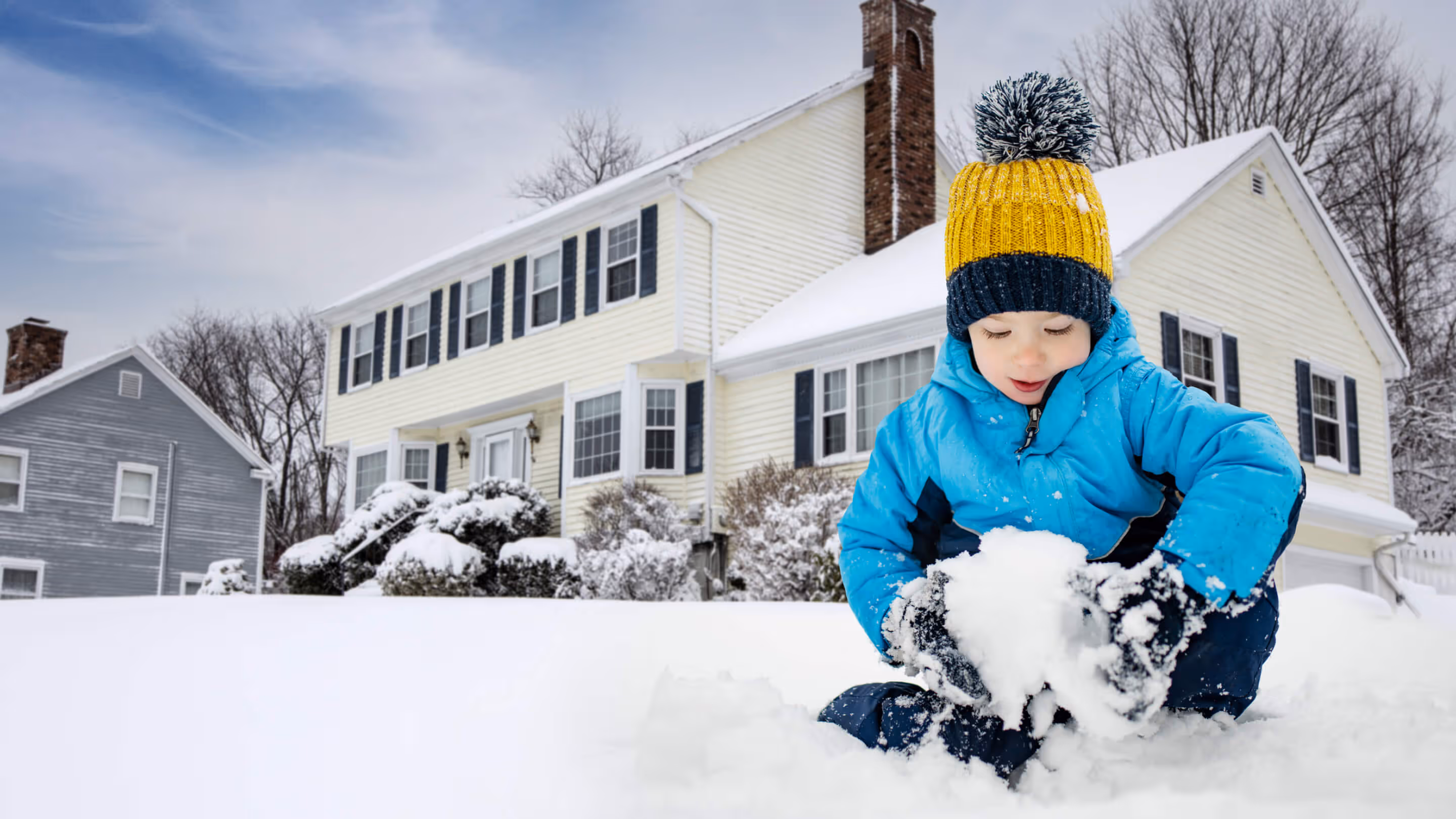 A child in a blue jacket and yellow hat plays with snow in front of a house on a snowy day.