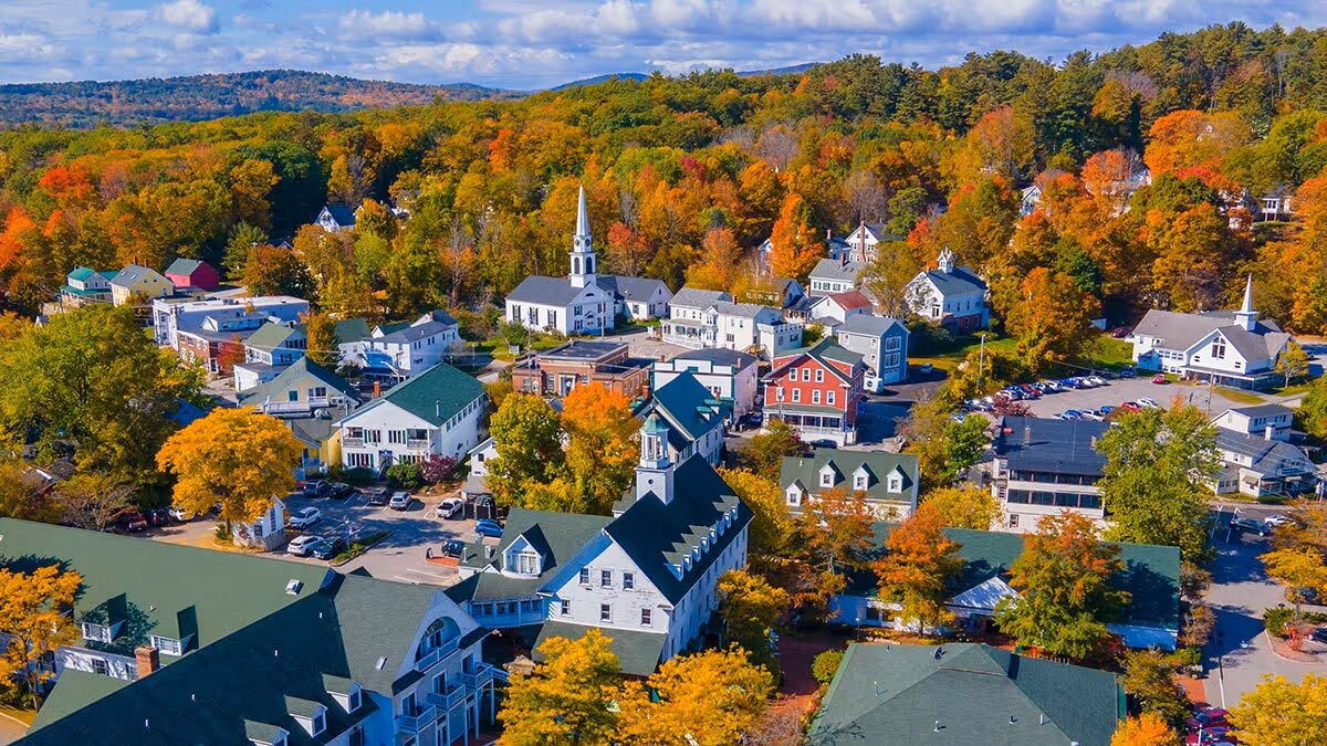Aerial view of a small town with colorful autumn trees and white church steeples under a partly cloudy sky.