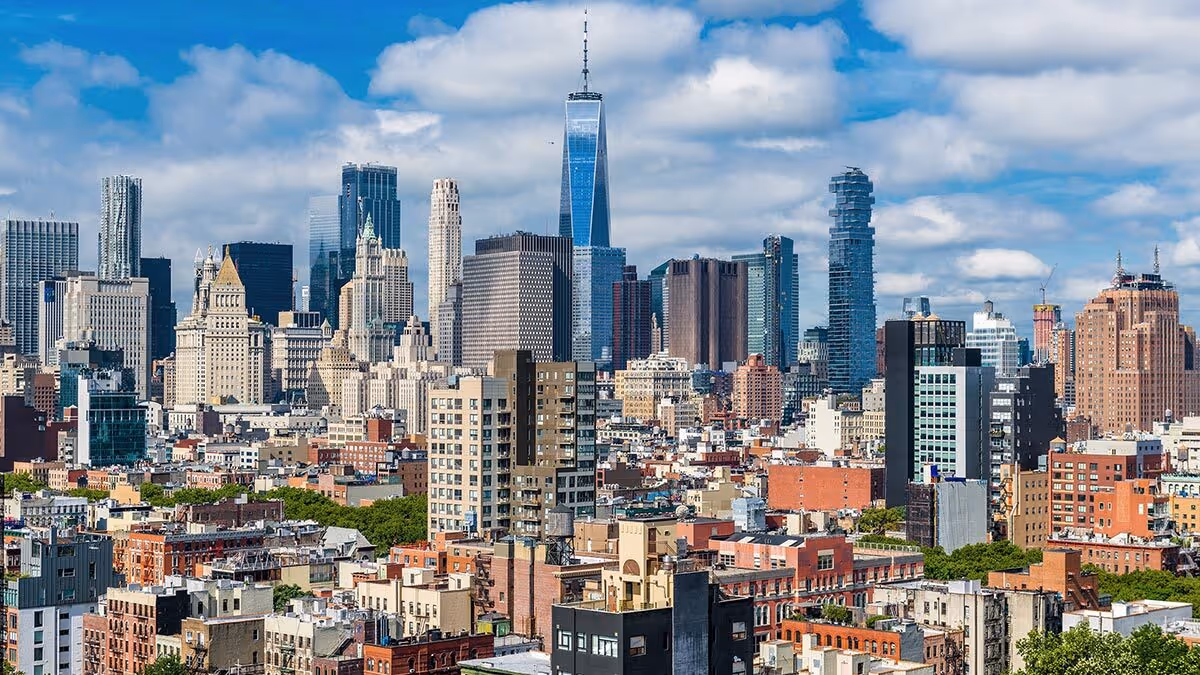 New York City skyline with tall skyscrapers, including One World Trade Center, under a partly cloudy sky.