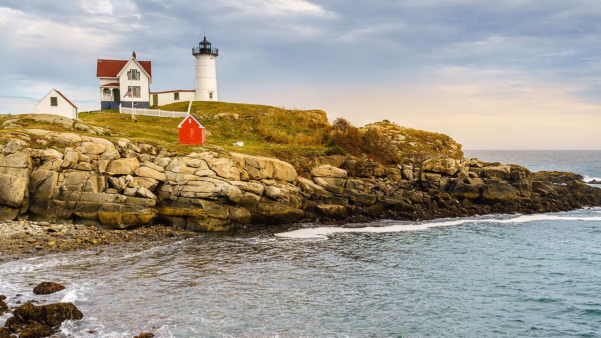 A white lighthouse and house sit on a rocky coastline above the ocean under a partly cloudy sky.