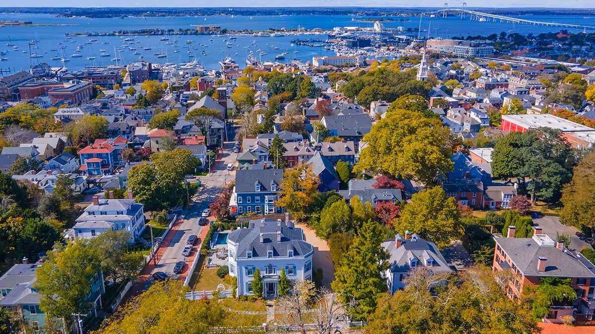 Aerial view of a coastal town with houses, trees, and a harbor filled with boats under a clear blue sky.