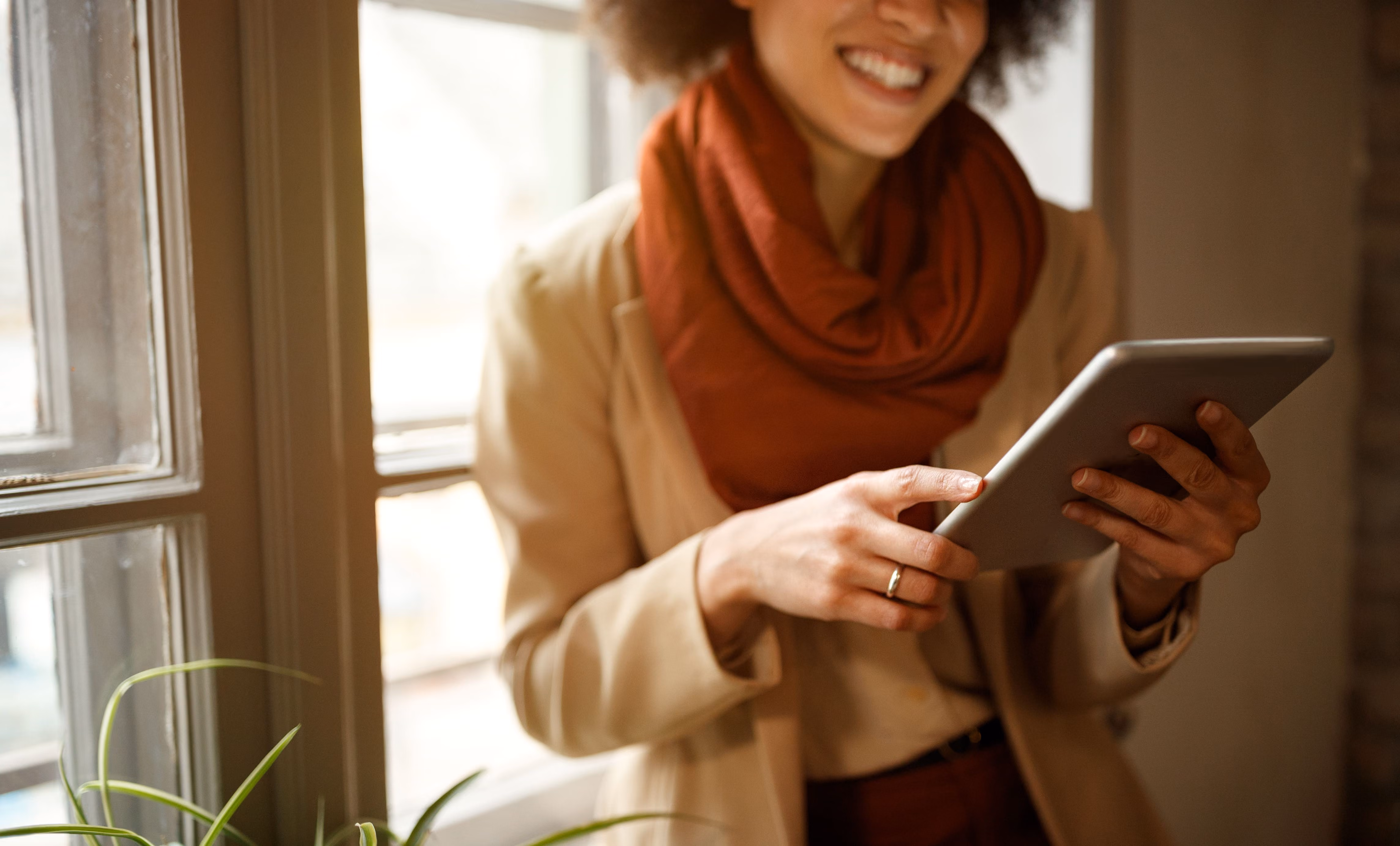 Smiling person wearing a beige coat and rust-colored scarf, using a tablet near a window with sunlight and a plant in the foreground.