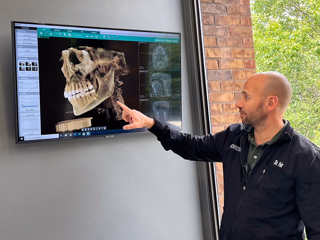 A man wearing a black jacket labeled Dr. Fair points at a large screen mounted on a wall displaying a 3D skull and dental X-rays.
