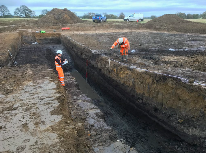 Two construction workers in orange safety gear and helmets are measuring and inspecting a deep excavation trench on a muddy site, with vehicles and soil piles in the background.