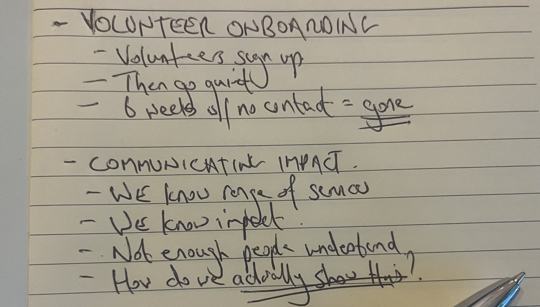 Handwritten notes on a notepad showing two rough problem statements - volunteer onboarding and communicating impact - with a pen resting on the page.