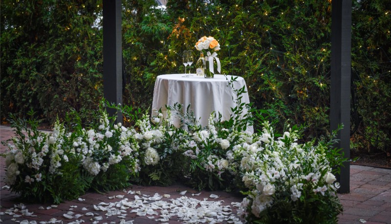 A bottle of champagne and flowers sit on a table with an arc of flowers in front of it and flower pedals on the ground