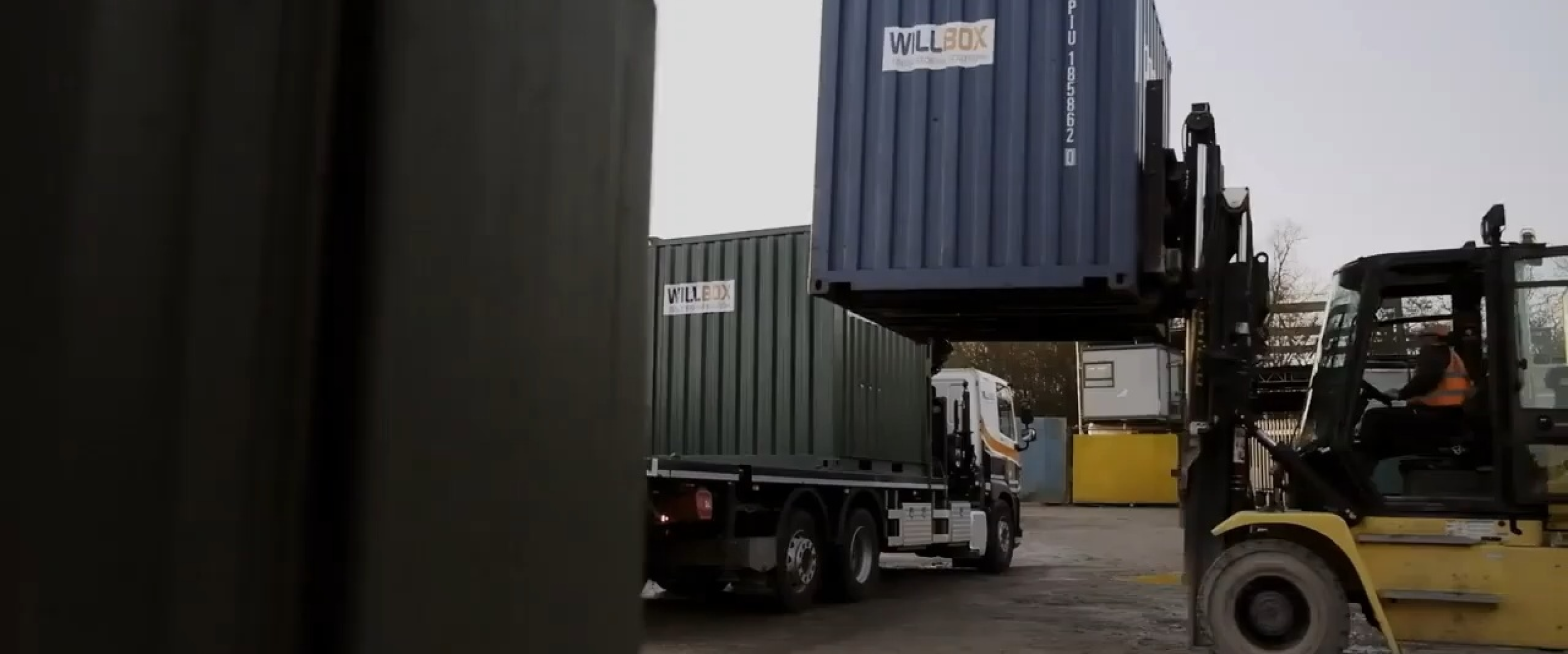 Forklift operator in orange vest lifting a blue WillBox shipping container near a truck with another green container.