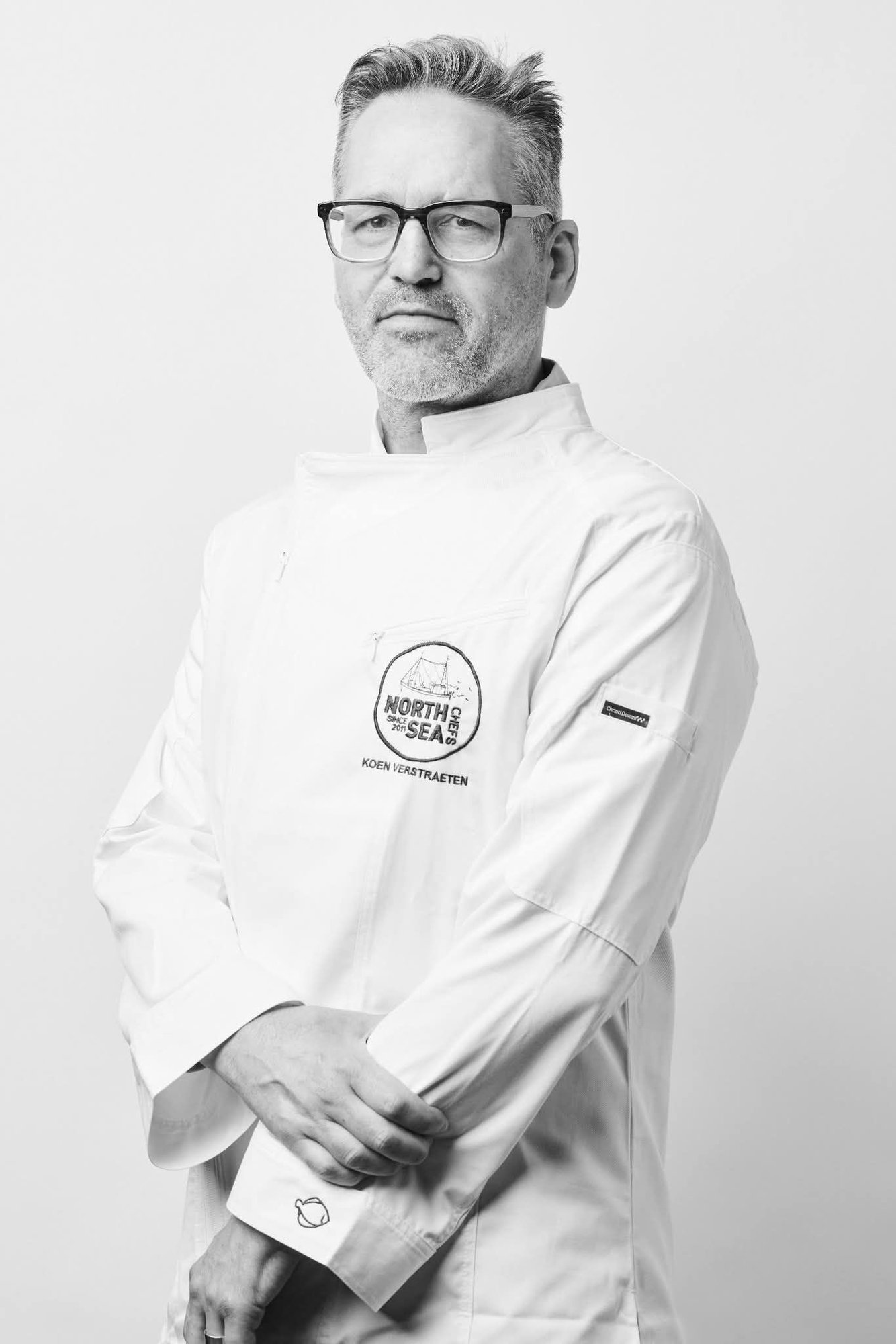 Portrait en noir et blanc du chef Koen Verstraeten en veste de cuisine blanche NorthSeaChefs, avec lunettes