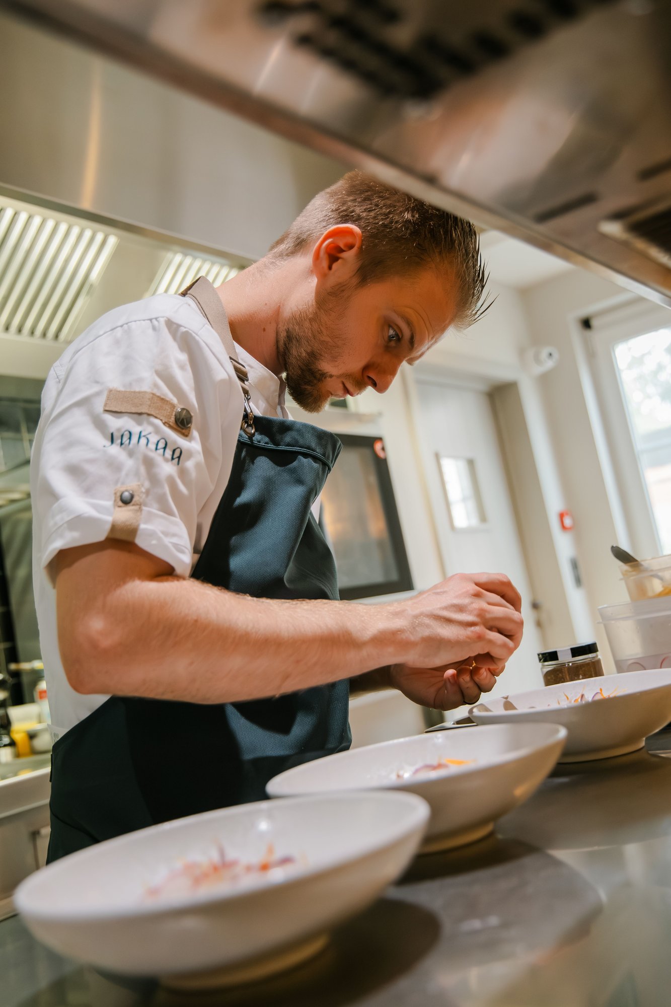 Photo couleur du chef Cedric Ongenae, en tablier foncé, finalisant des assiettes dans la cuisine de Jakaa