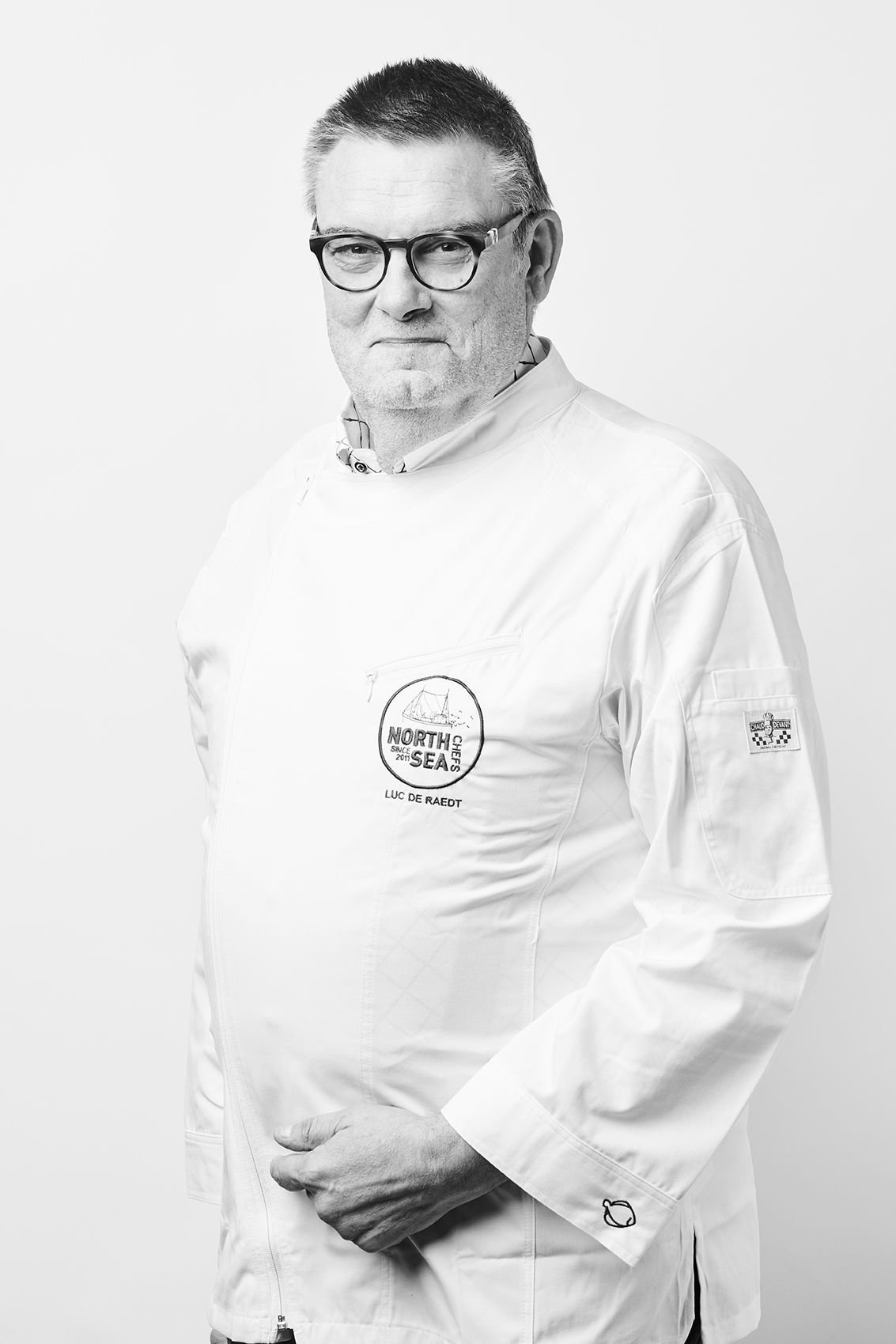 Portrait en noir et blanc du chef Luc De Raedt en veste de cuisine blanche NorthSeaChefs, avec lunettes