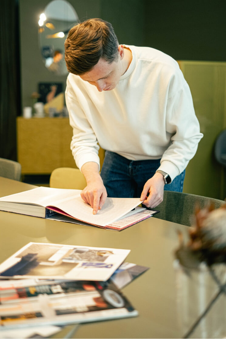 Young man in white sweater leaning over table, pointing at pages in an open book in a cozy room.