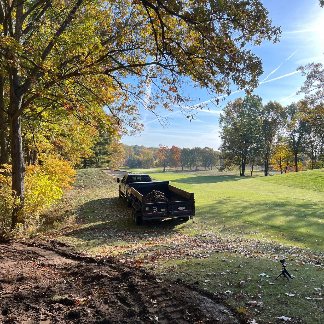 Black truck with trailer parked on green grass beside trees with autumn leaves under a clear blue sky.