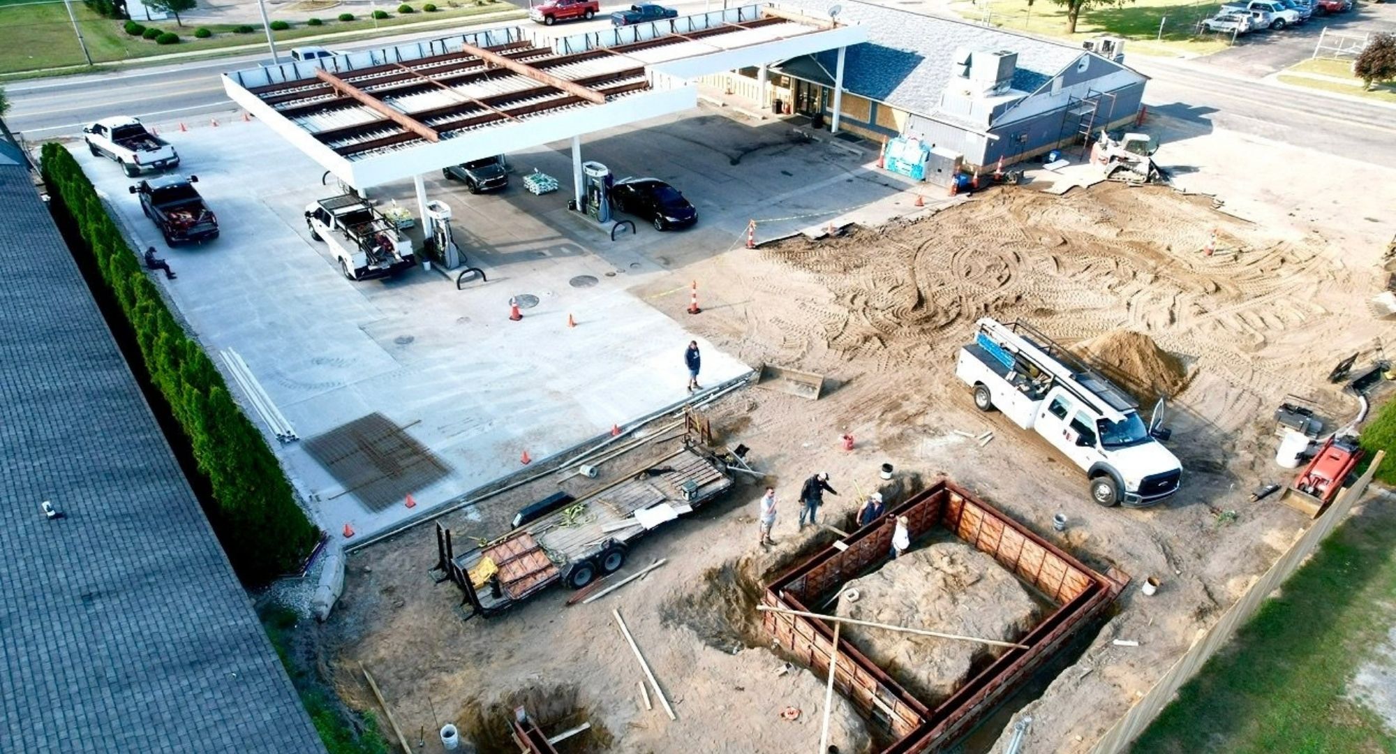 Construction site next to gas station with trucks, workers, dirt excavation, and building materials.