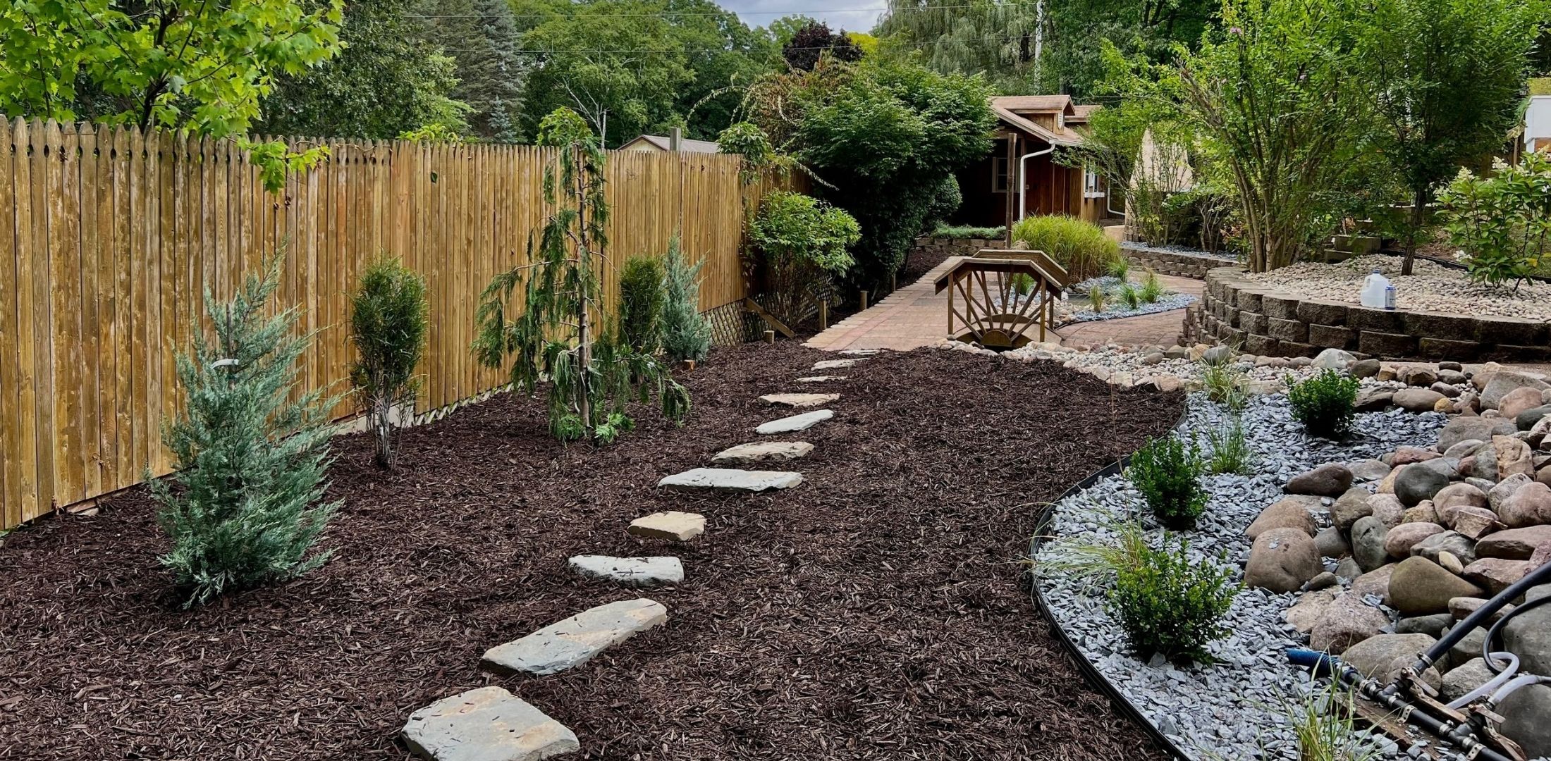 Backyard landscaping with mulch, stepping stones, shrubs, rock garden, and wooden fence in daytime.