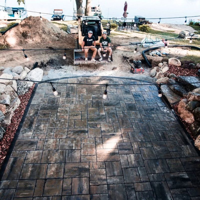 Two men sitting on a construction vehicle behind a newly paved stone patio with string lights overhead.
