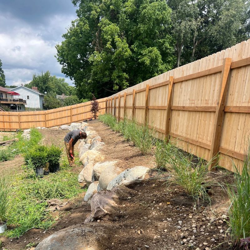 Person working near large rocks along a newly planted garden bed with a tall wooden fence.