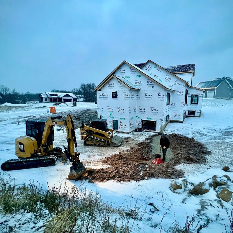 Snowy construction site with house wrapped in Tyvek and workers using heavy machinery and tools.